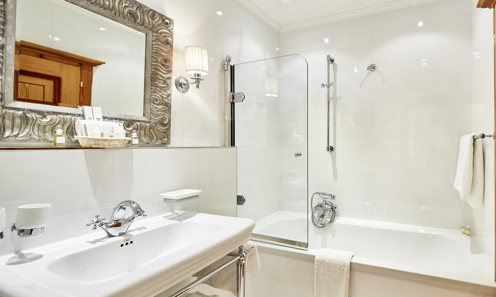 Elegantes weißes Badezimmer mit Waschbecken, Spiegel, Glastür zur Dusche, Badewanne und Handtüchern an der Wand.