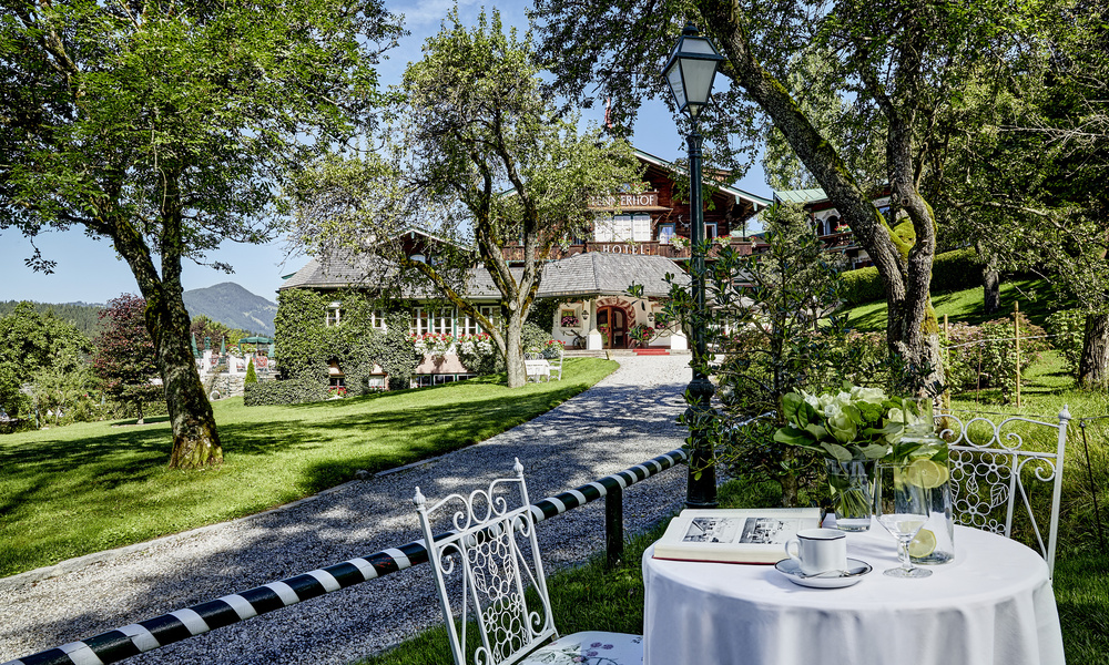 Ein für eine Person gedeckter Tisch blickt auf einen Gartenweg, der zu einer rustikalen, von Bäumen und Grün umgebenen Hütte führt.