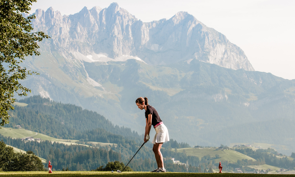 Eine Frau bereitet sich darauf vor, einen Golfschläger auf einem Grün mit Bergen im Hintergrund zu schwingen.