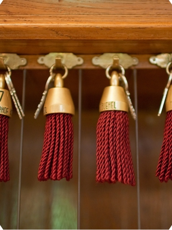 Vier alte Hotelschlüssel mit roten Quasten hängen an Haken vor einem hölzernen Hintergrund.