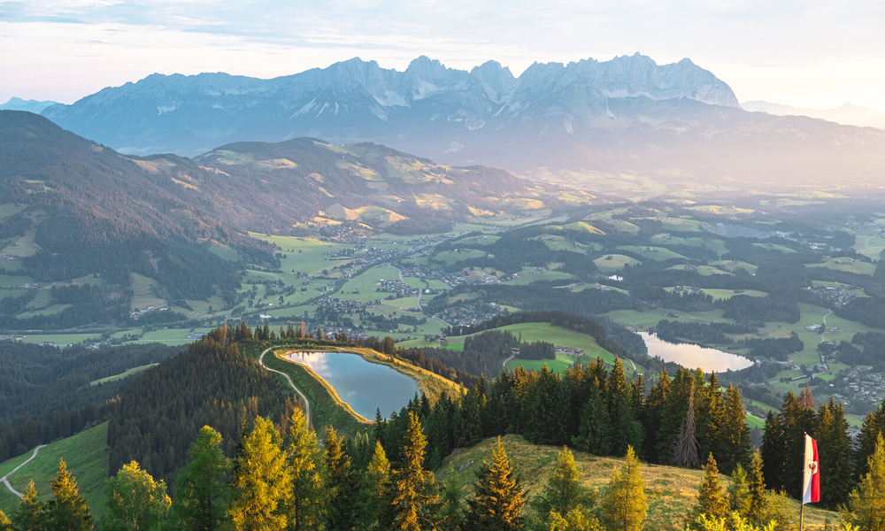 Malerische Berglandschaft mit Wäldern, Seen, Dörfern und entfernten Gipfeln im sanften Morgenlicht.
