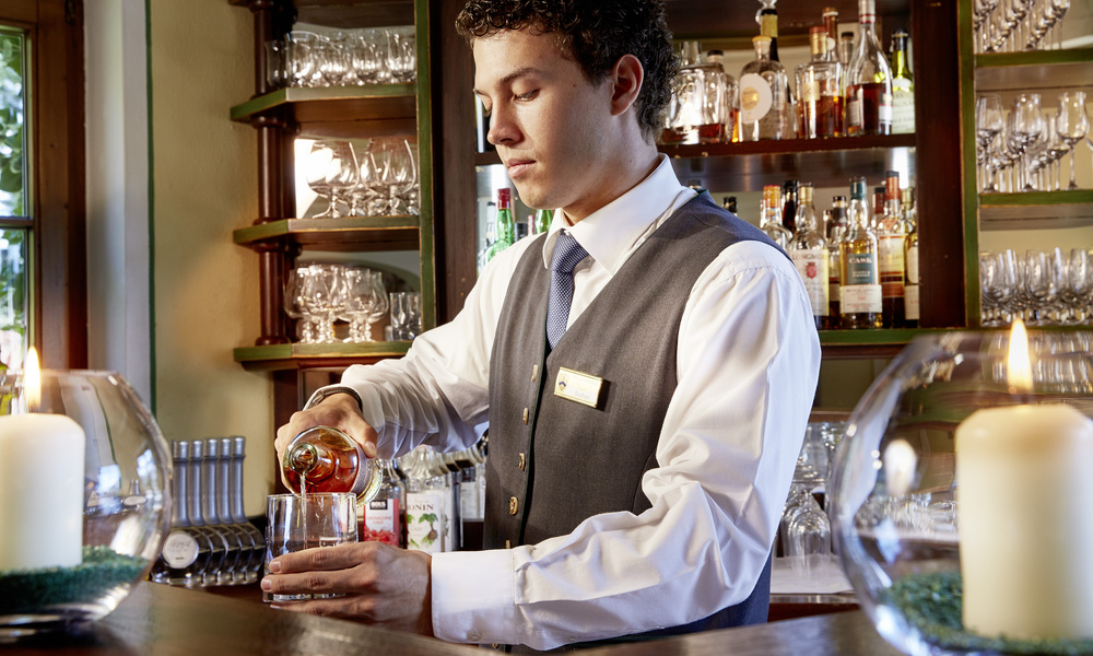 Ein Barkeeper in Weste gießt einen Drink in ein Glas hinter einer gut bestückten Bar mit Gläsern und Flaschen.
