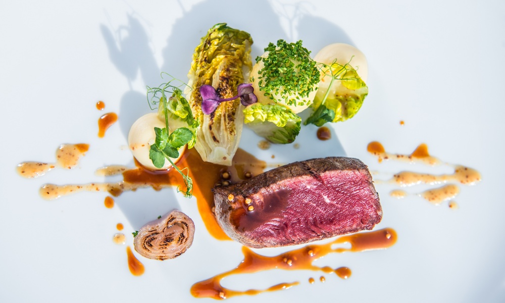 Ein plattiertes Gourmet-Menü mit blutigem Steak, Grünzeug, Soßenträufeln und kunstvoll arrangiertem Gemüse auf einem weißen Teller.