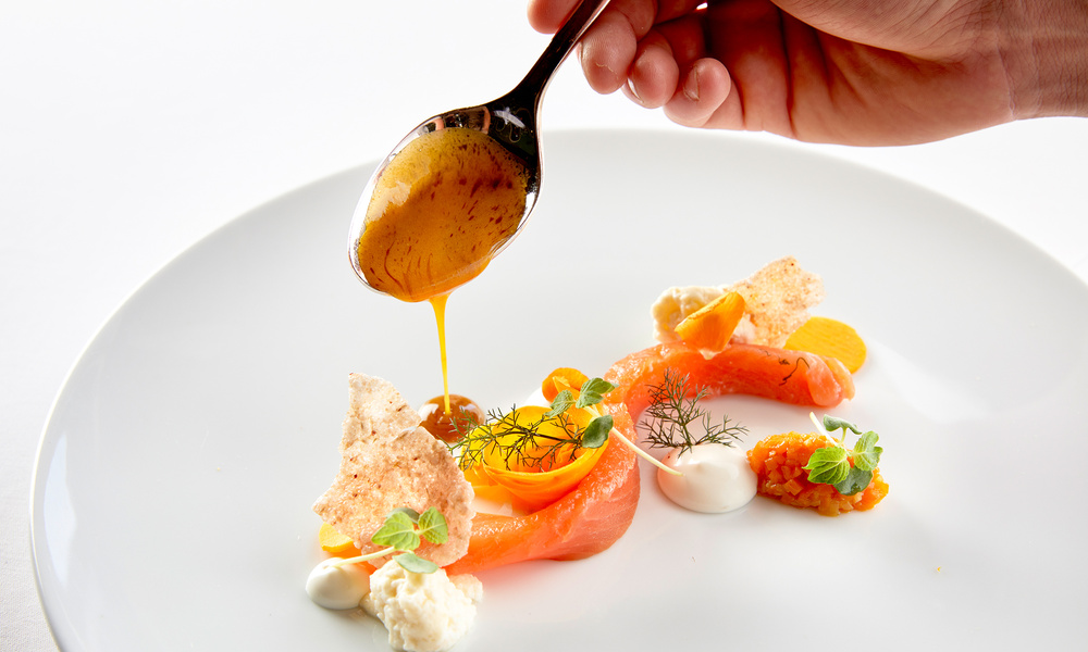 Eine Hand löffelt Sauce auf ein Gourmetgericht mit Lachs, Kräutern, Saucen und Garnierungen auf einem weißen Teller.
