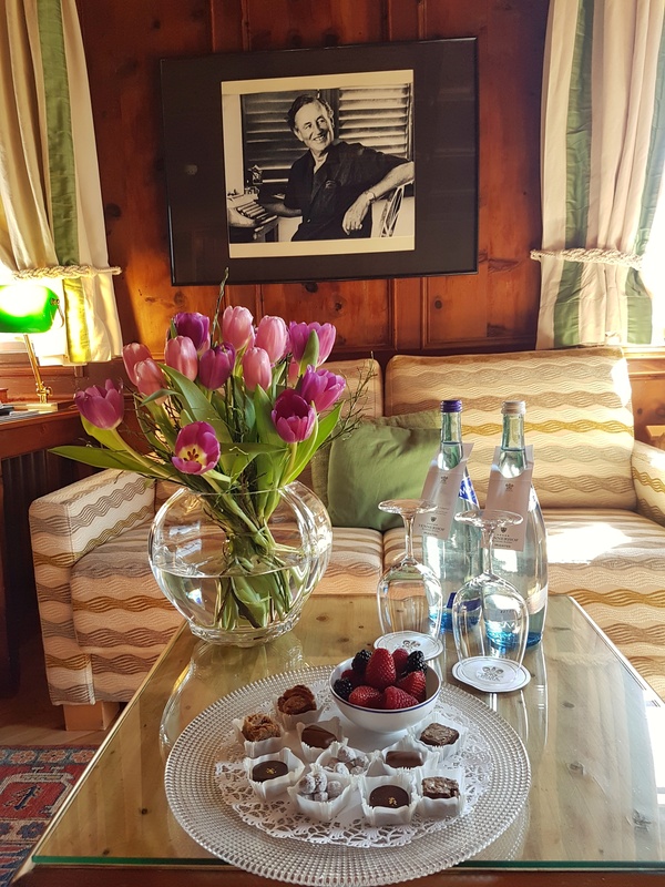 Vase mit lila Tulpen, Snacks und Wasserflaschen auf einem Glastisch, dahinter ein Schwarz-Weiß-Porträt.