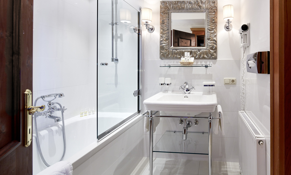 Modernes weißes Bad mit Badewanne, Waschbecken, verziertem Spiegel, Glasablagen und Wandleuchten.