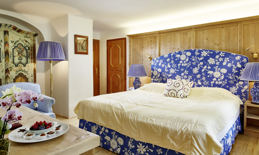 Gemütliches Schlafzimmer mit blauem, geblümtem Kopfteil, gelber Bettwäsche, zwei blauen Lampen und einem Teller mit Obst auf dem Tisch.