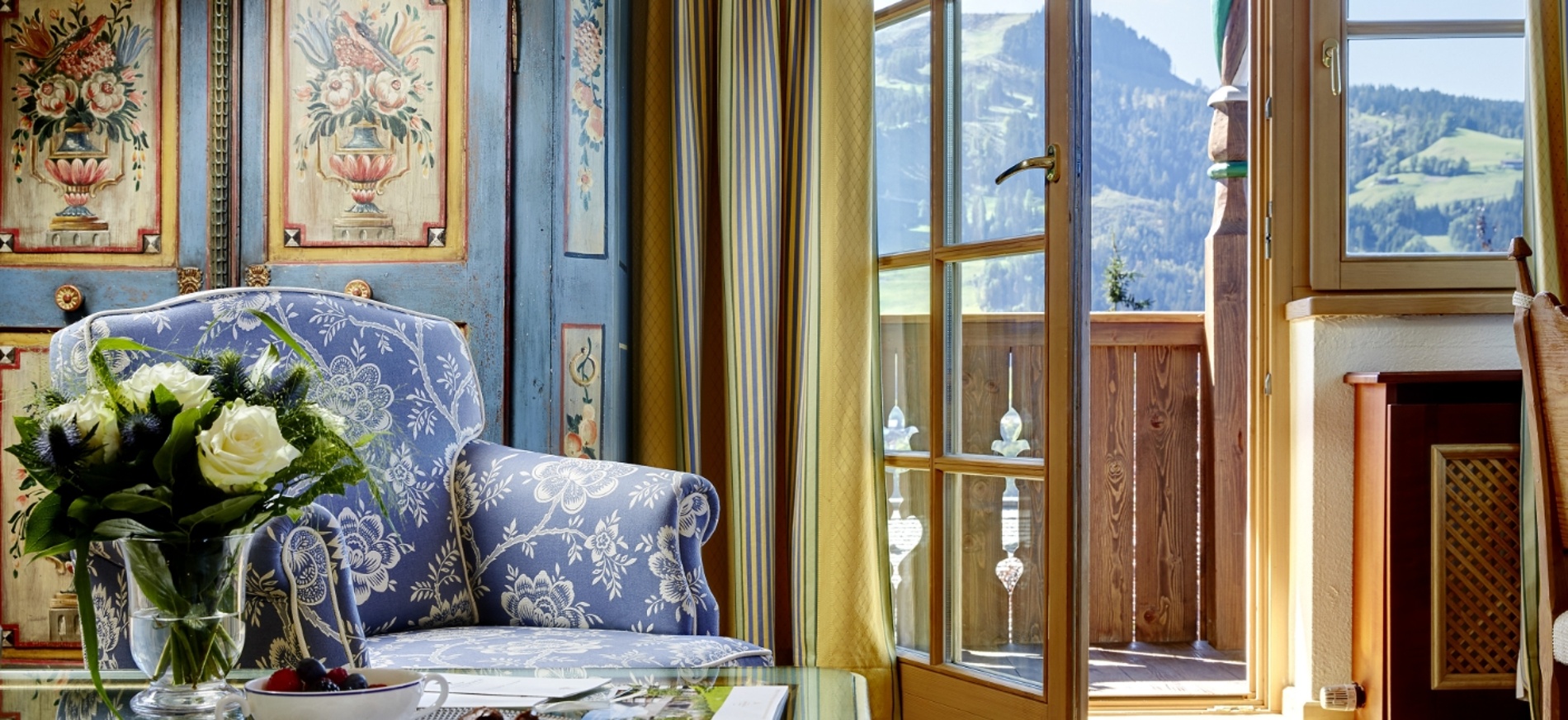 Ein gemütlicher, geblümter Sessel neben einer Glastür führt auf einen Balkon mit Blick auf die Berge.