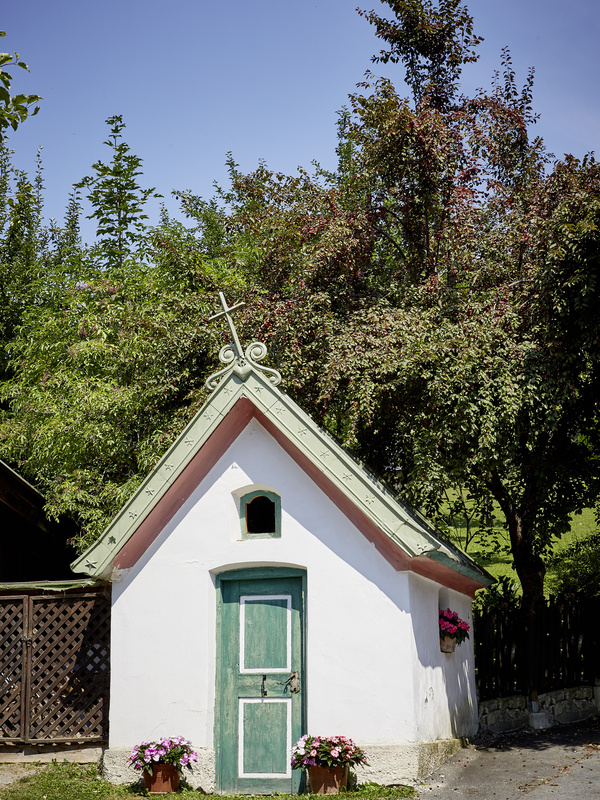 Kleine weiße Kapelle mit grüner Tür, Kreuz auf dem Dach und Blumen in Töpfen; Bäume im Hintergrund.