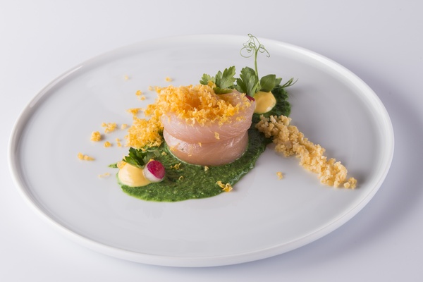 Ein Gourmetgericht mit dünnen Fischscheiben, grüner Soße, gelben Bröseln und kleinen Kräutern auf einem weißen Teller.