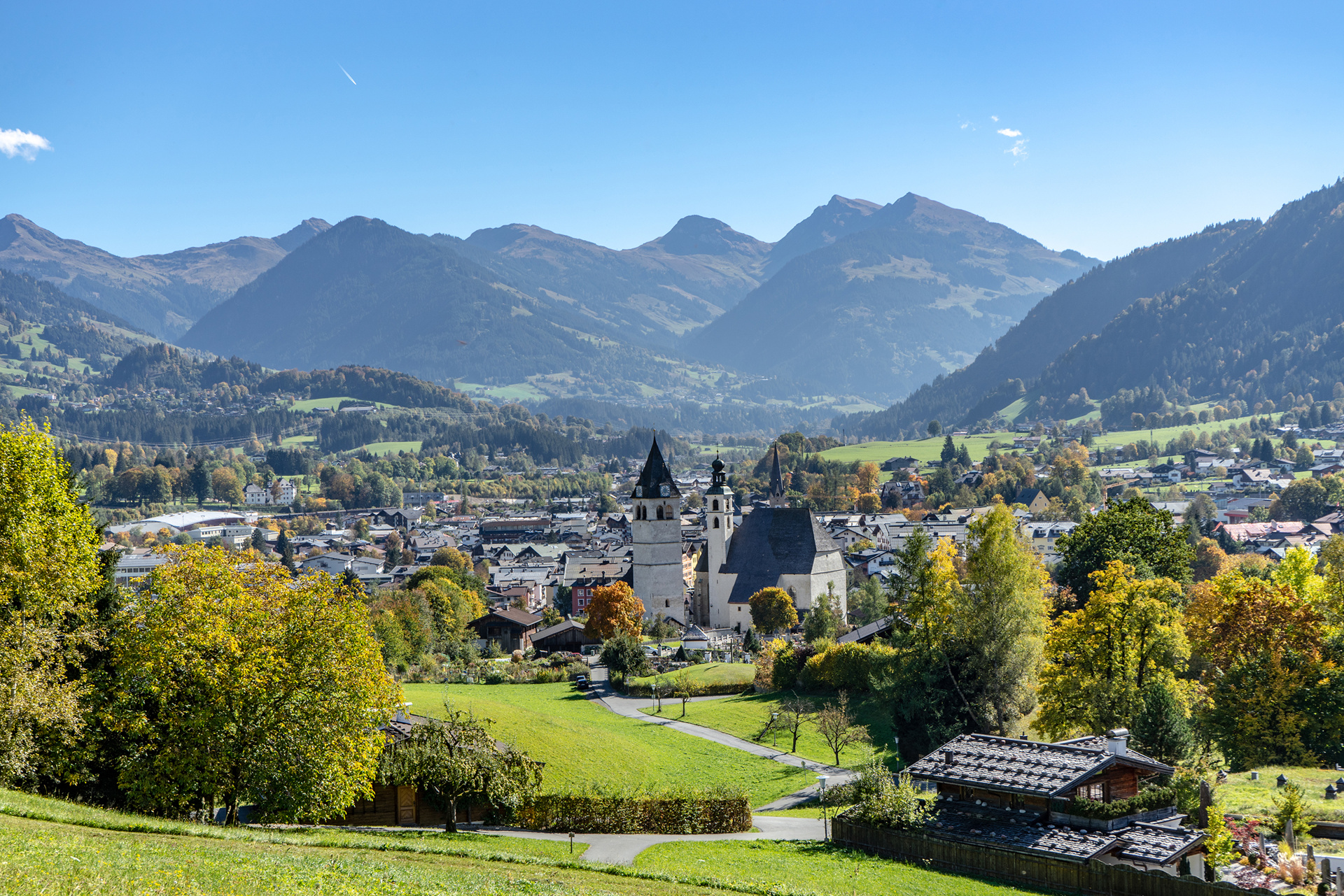 Ein malerisches Dorf mit einem Kirchturm, grünen Feldern und Bergen im Hintergrund unter einem klaren blauen Himmel.