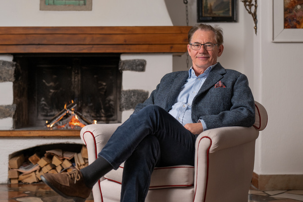 Ein lächelnder Mann mit Brille sitzt in einem Sessel vor einem beleuchteten Kamin in einem gemütlichen, mit Kunst gefüllten Raum.
