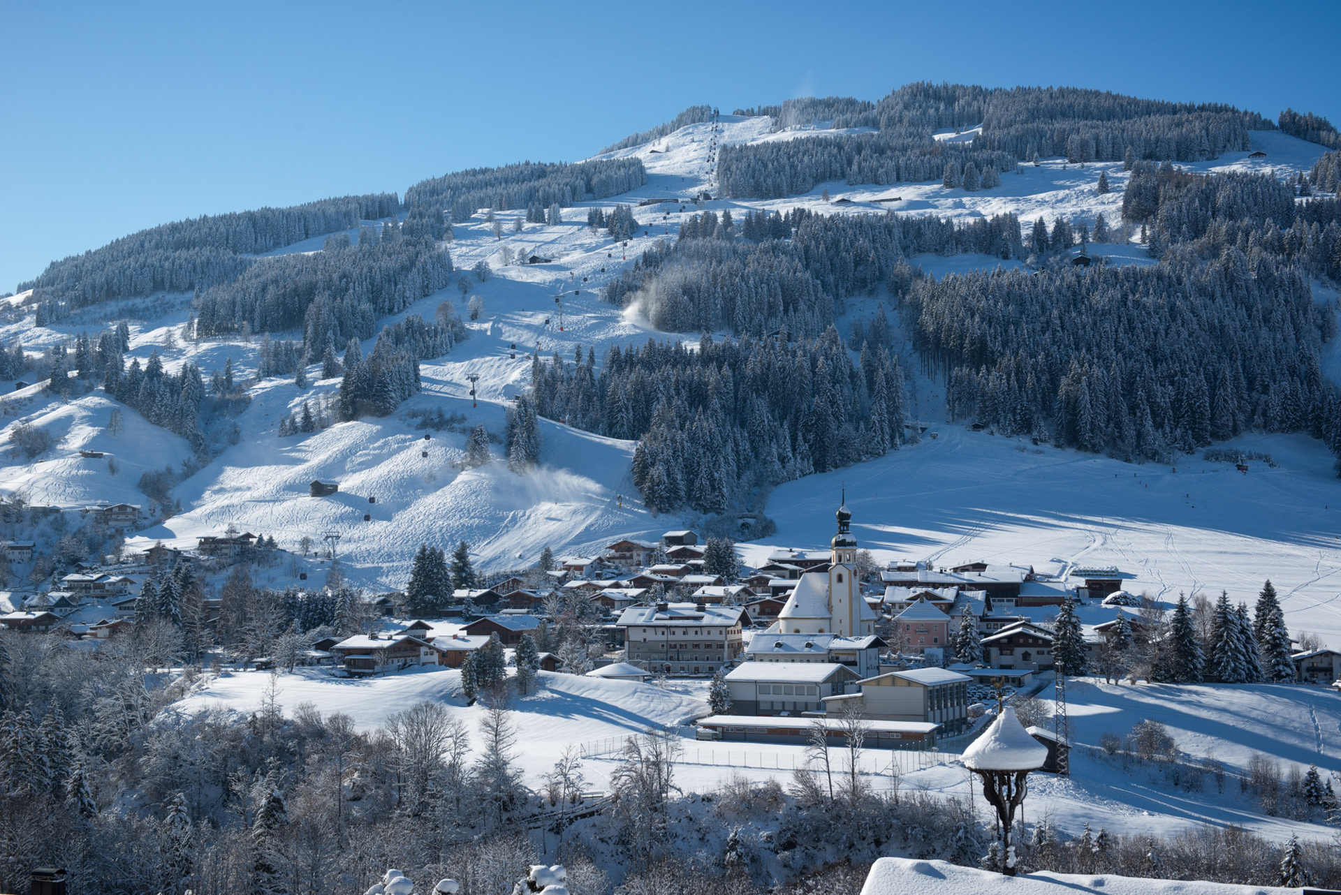 Ein verschneites Dorf mit einer Kirche am Fuße eines bewaldeten, verschneiten Berges unter blauem Himmel.