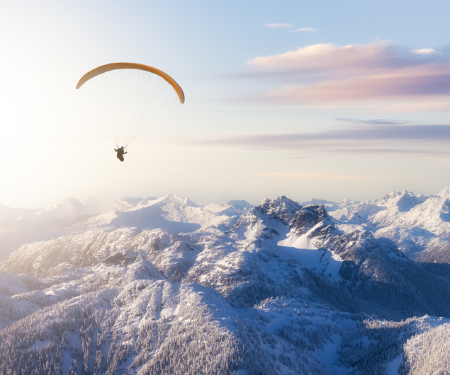 Ein Gleitschirmflieger schwebt über schneebedeckten Berggipfeln unter einem hellen Himmel mit weichen Wolken bei Sonnenuntergang.