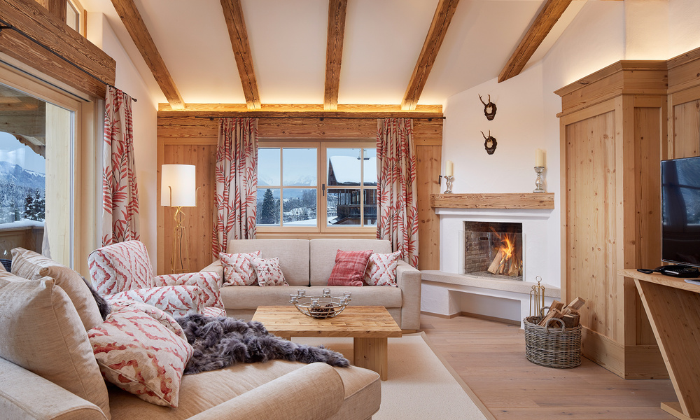 Gemütliches Wohnzimmer mit beigen Sofas, rot gemusterten Kissen, Kamin, Holzbalken und Bergblick durch das Fenster.