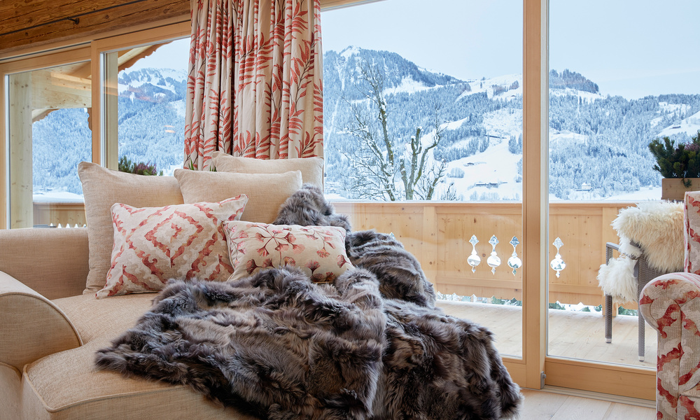 Gemütlicher Loungesessel mit Fellüberwurf, Kissen und Blick auf die verschneiten Berge durch große Glastüren.