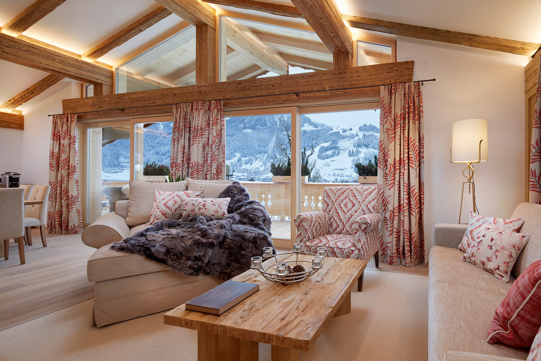 Warm, cozy living room with wooden beams, large windows, patterned curtains and mountain views.