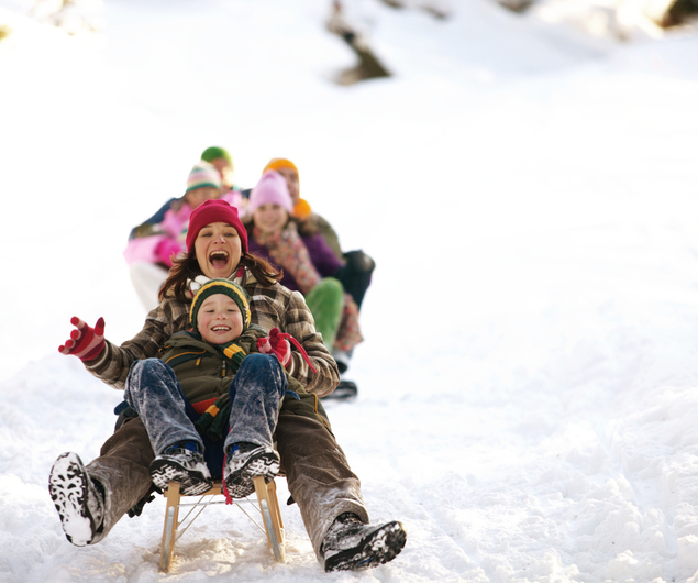 Eine Gruppe von Menschen, angeführt von einer Frau und einem Kind, fährt mit dem Schlitten einen verschneiten Hügel hinunter und lächelt fröhlich.