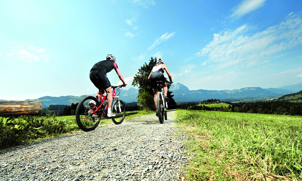 Zwei Radfahrer fahren auf einem Schotterweg durch eine grasbewachsene Landschaft mit Bergen im Hintergrund.