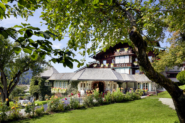 Charmantes Alpenhotel, umgeben von Bäumen, Blumen und grünen Rasenflächen unter einem klaren blauen Himmel.