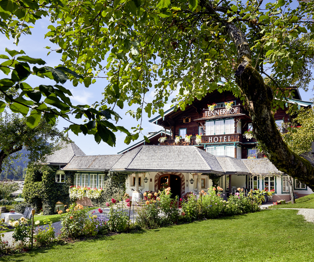 Charmantes Alpenhotel, umgeben von Bäumen, Blumen und grünen Rasenflächen unter einem klaren blauen Himmel.