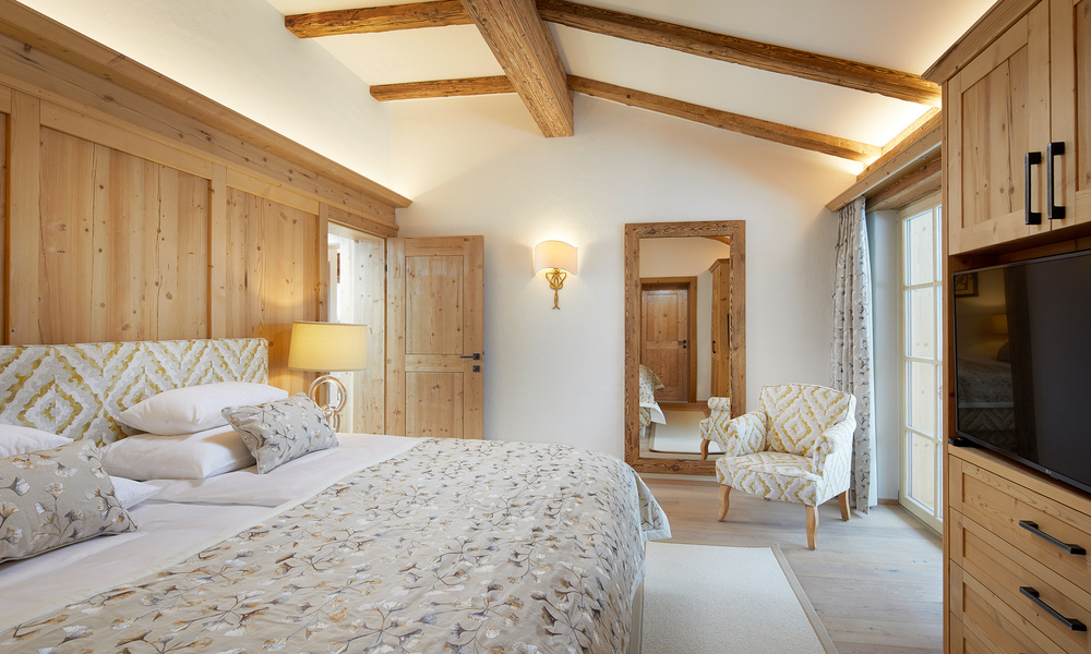 Gemütliches Schlafzimmer mit Holzwänden, gemustertem Bettzeug, Sessel, großem Spiegel, TV und sichtbaren Deckenbalken.