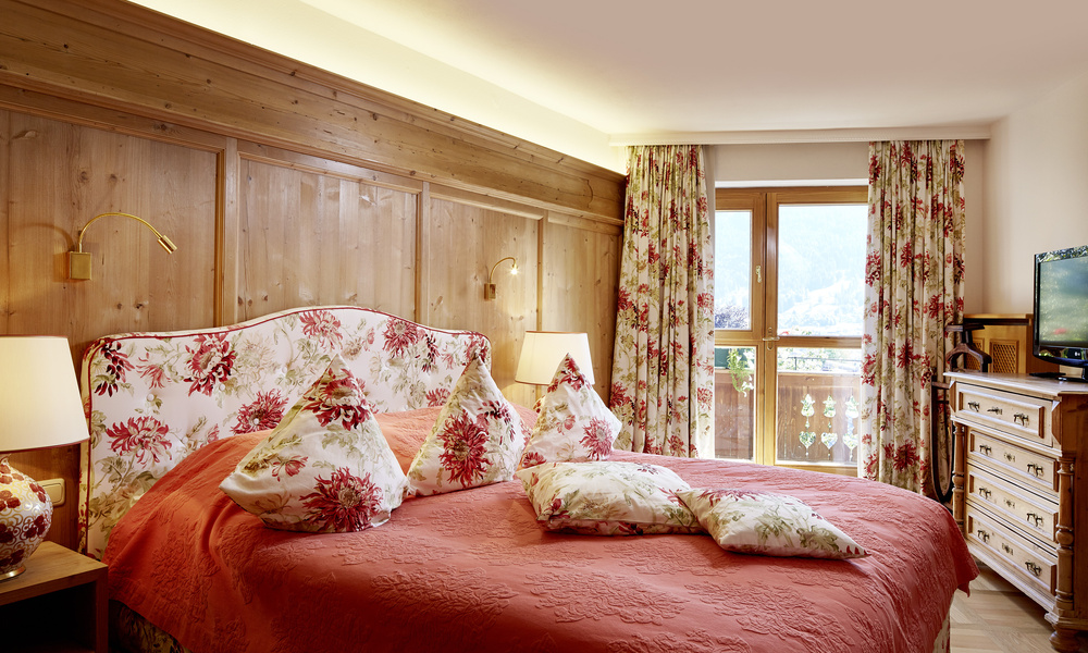 Ein gemütliches Schlafzimmer mit geblümtem Bettzeug, Holzwänden und einem Fenster mit Blick auf einen malerischen Balkon.