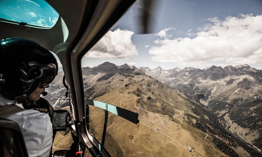 Ein Pilot fliegt mit einem Hubschrauber über weite, zerklüftete Berge unter einem teilweise bewölkten Himmel.