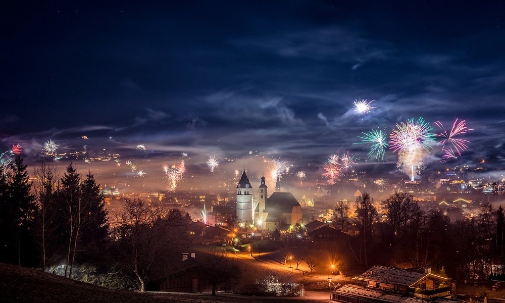 Ein Feuerwerk erhellt den Nachthimmel über einer Stadt mit einer Kirche und Häusern, umgeben von Bäumen und Nebel.