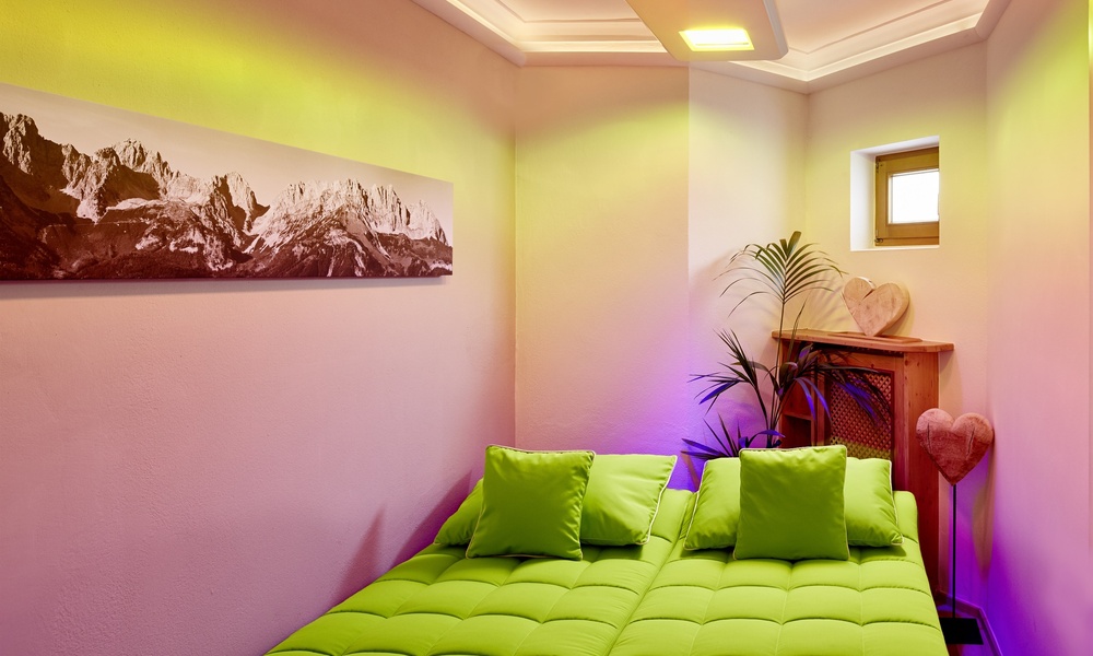 Ein gemütliches Schlafzimmer mit einem hellgrünen Bett, Wandkunst aus den Bergen, Pflanzen und sanfter violetter und gelber Beleuchtung.