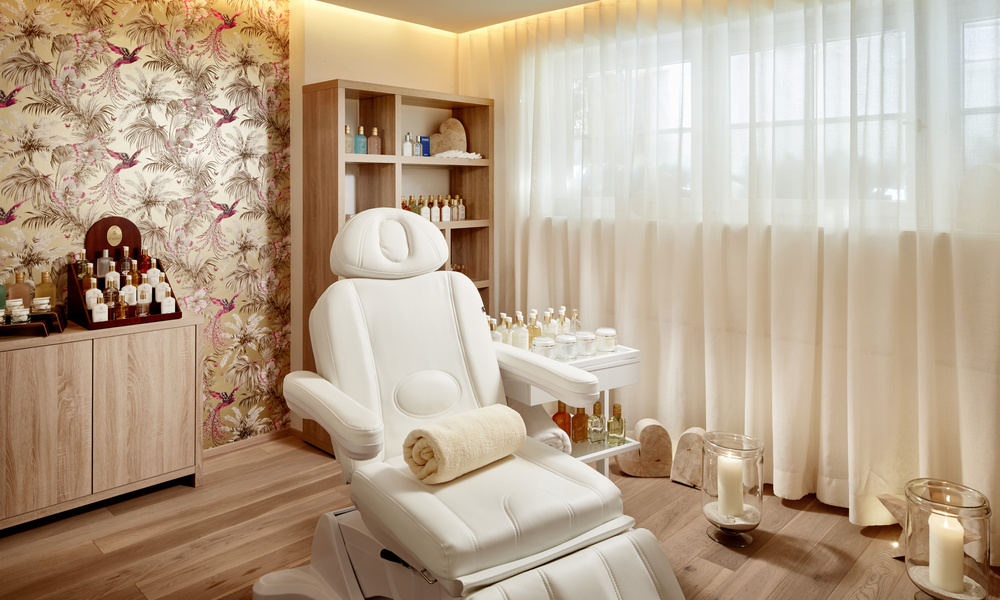 Ein moderner Spa-Raum mit weißem Behandlungsstuhl, gerolltem Handtuch, Kerzen und Regalen mit Hautpflegeprodukten.
