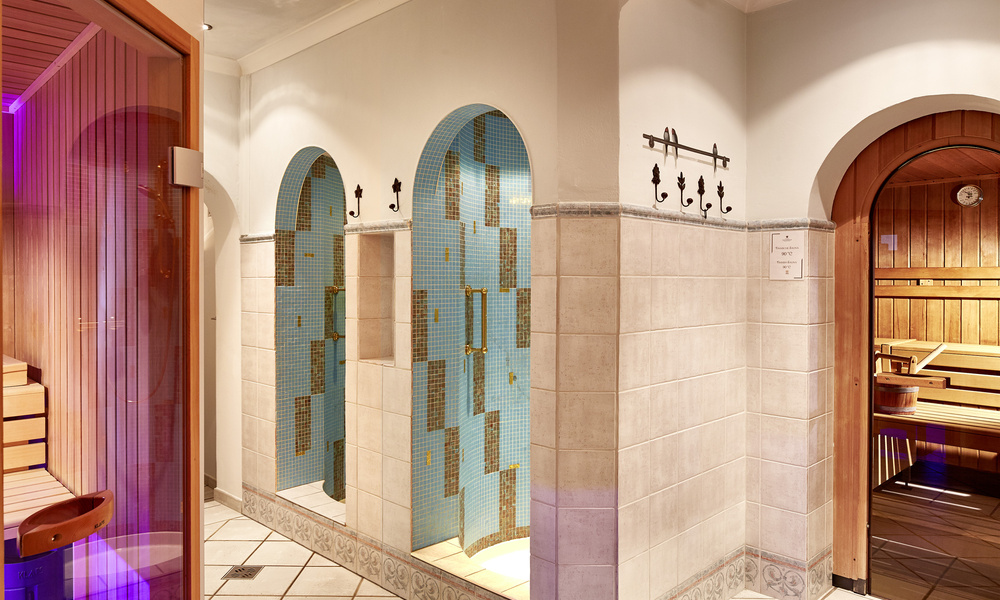Gefliester Sauna- und Duschbereich mit Holzbänken, gewölbten Türöffnungen und Wandhaken.