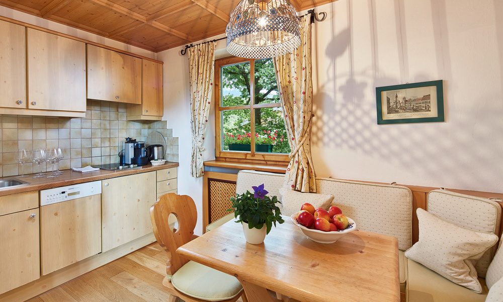 Gemütliche Küche mit hellen Holzschränken, einem Esstisch, einer Obstschale und einem Fenster mit Blick ins Grüne.