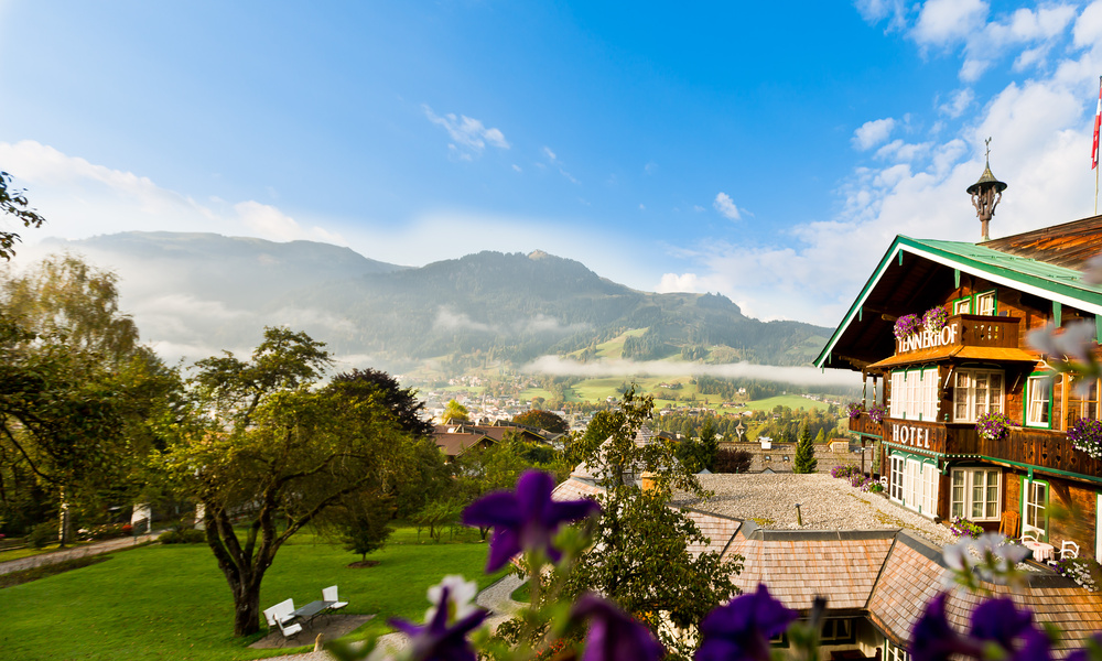 Blick auf ein Hotel und einen Garten mit Bergen und tief hängenden Wolken im Hintergrund an einem klaren Tag.