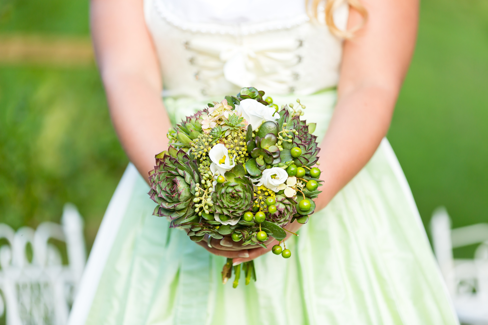 A woman in a light green dress holds a bouquet of succulents and white flowers outside.
