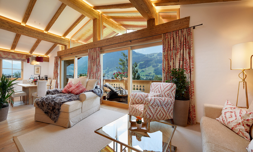 Gemütliches Wohnzimmer mit Holzbalken, gemusterten Stühlen, Blick auf die Berge und großen Fenstern, die viel Tageslicht hereinlassen.