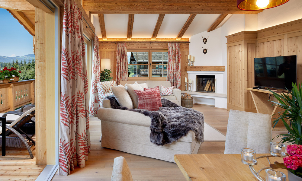 Gemütliches Wohnzimmer mit Holzbalken, großen Fenstern, Kamin und einem mit Kissen und einer Felldecke bedeckten Sofa.