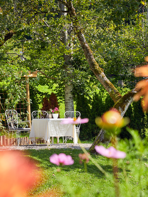 Weißer Metalltisch und Stühle mit Blumen in einem grünen, sonnendurchfluteten Garten, umgeben von blühenden Pflanzen und Bäumen.