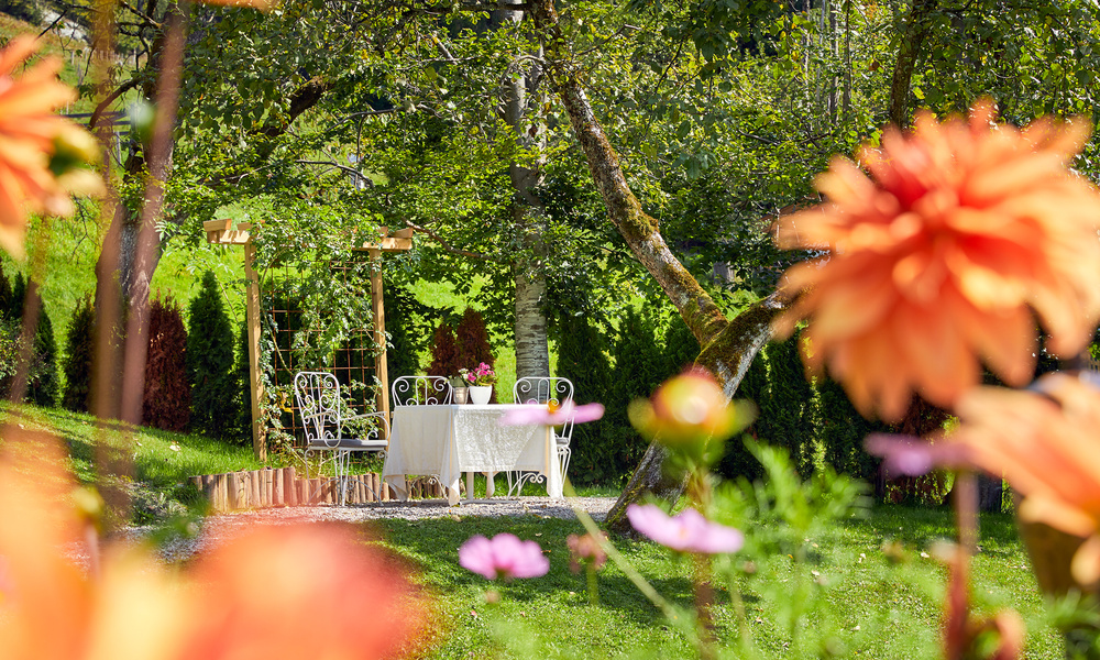 Weißer Metalltisch und Stühle unter Bäumen in einem sonnigen Garten, mit orangefarbenen Blumen im Vordergrund.