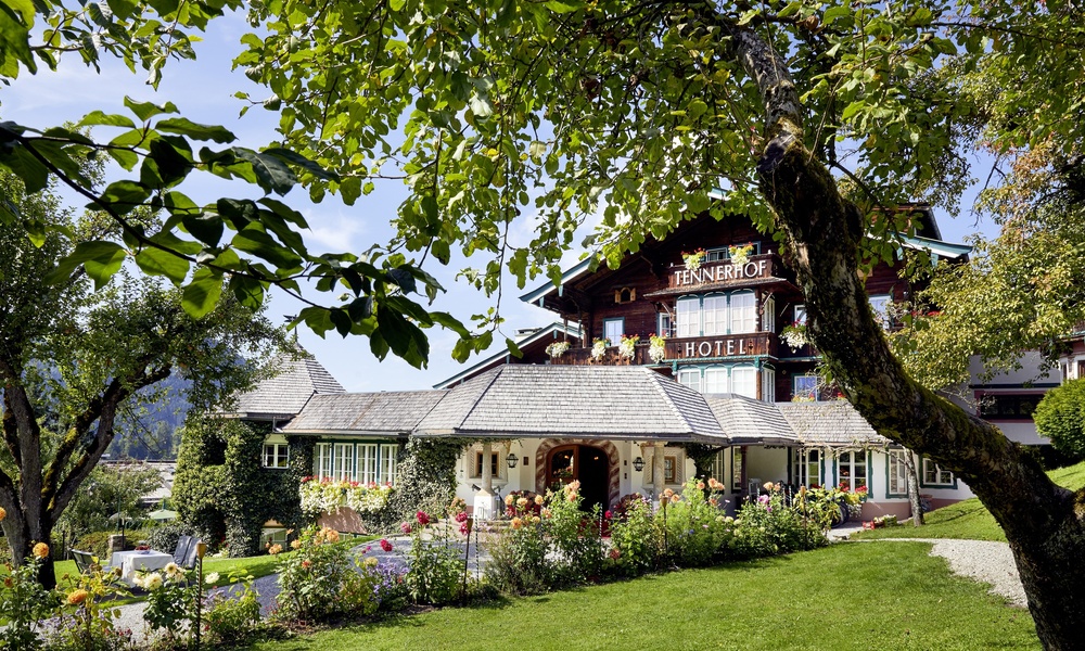 Charmantes Alpenhotel, umgeben von üppigen Gärten und Bäumen unter blauem Himmel.