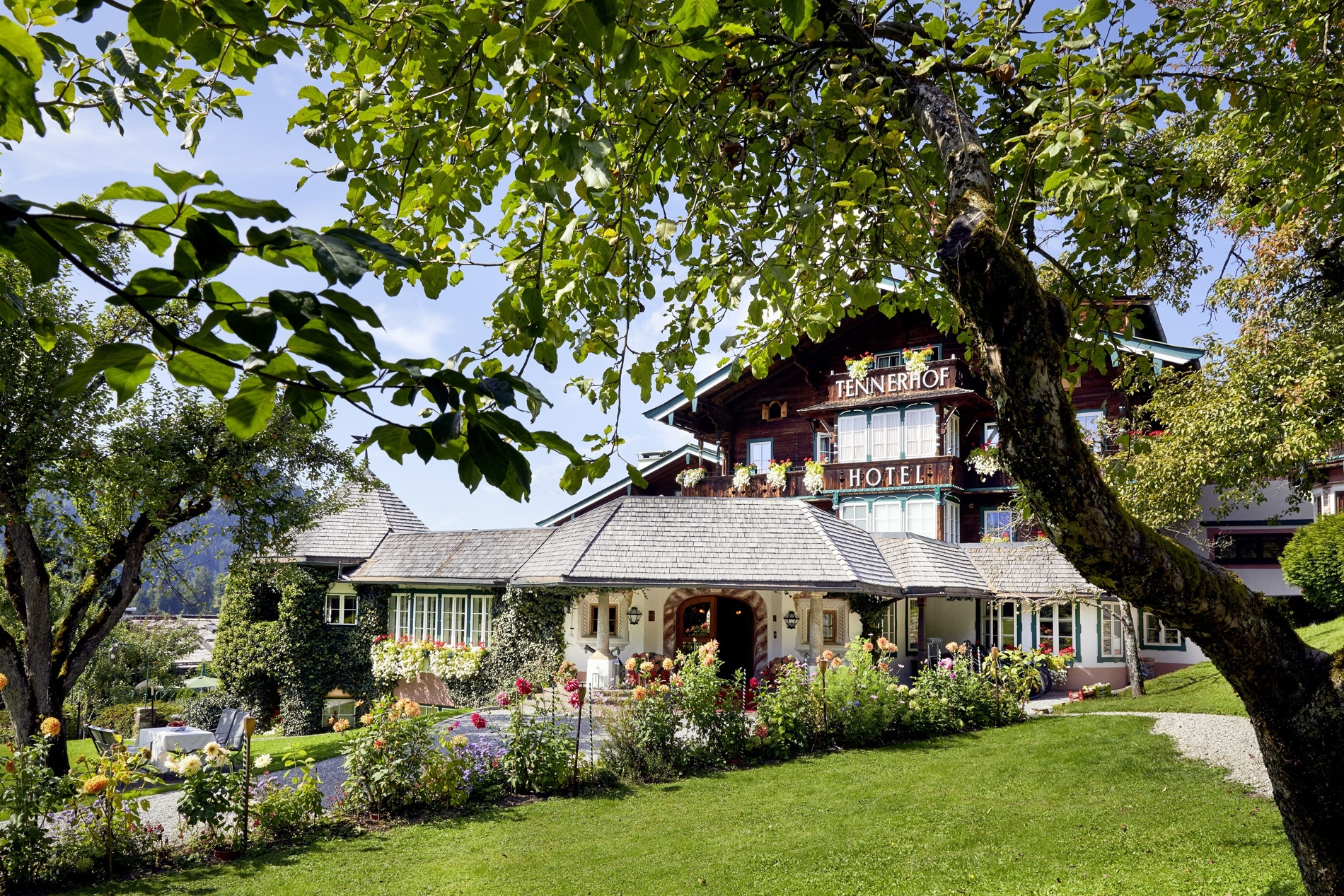 Charmantes Alpenhotel, umgeben von üppigen Gärten und Bäumen unter blauem Himmel.