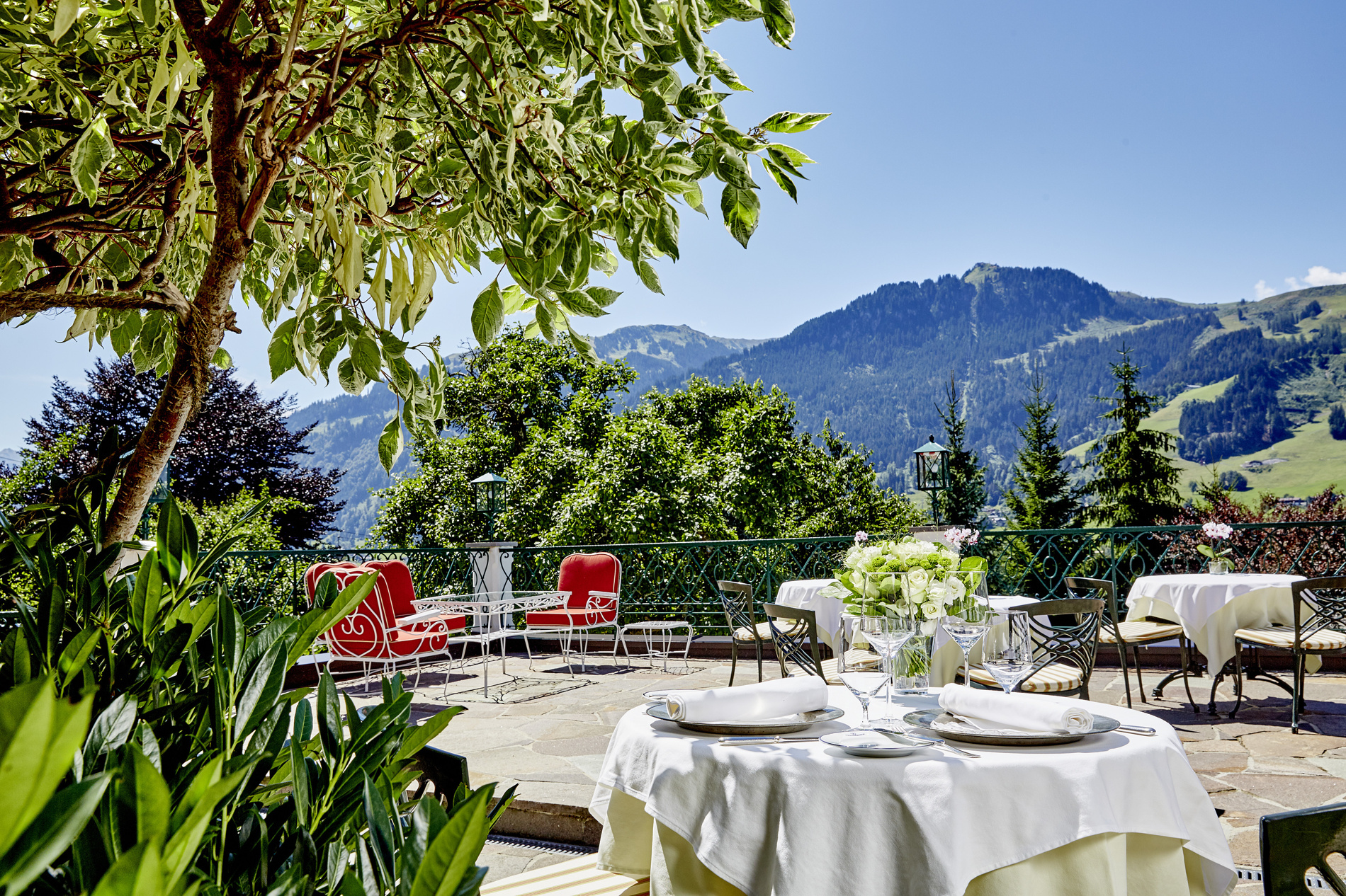 Restaurant terrace with white tablecloths, red chairs and a view of the mountains under a bright blue sky.