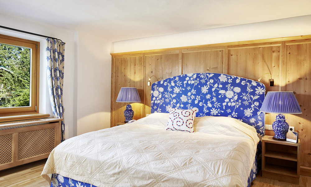 Gemütliches Schlafzimmer mit einem blauen geblümten Kopfteil, zwei blauen Lampen und cremefarbener Bettwäsche neben einem Fenster.