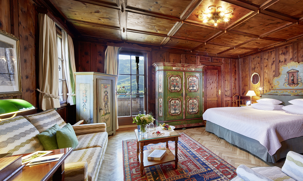 Gemütliches, rustikales Schlafzimmer mit Holzvertäfelung, bemaltem Kleiderschrank, Doppelbett, Sofa und Bergblick durch das Fenster.