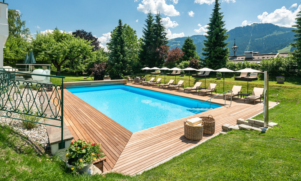 Rechteckiger Außenpool mit Liegestühlen und Sonnenschirmen, umgeben von Bäumen und Bergen unter einem blauen Himmel.
