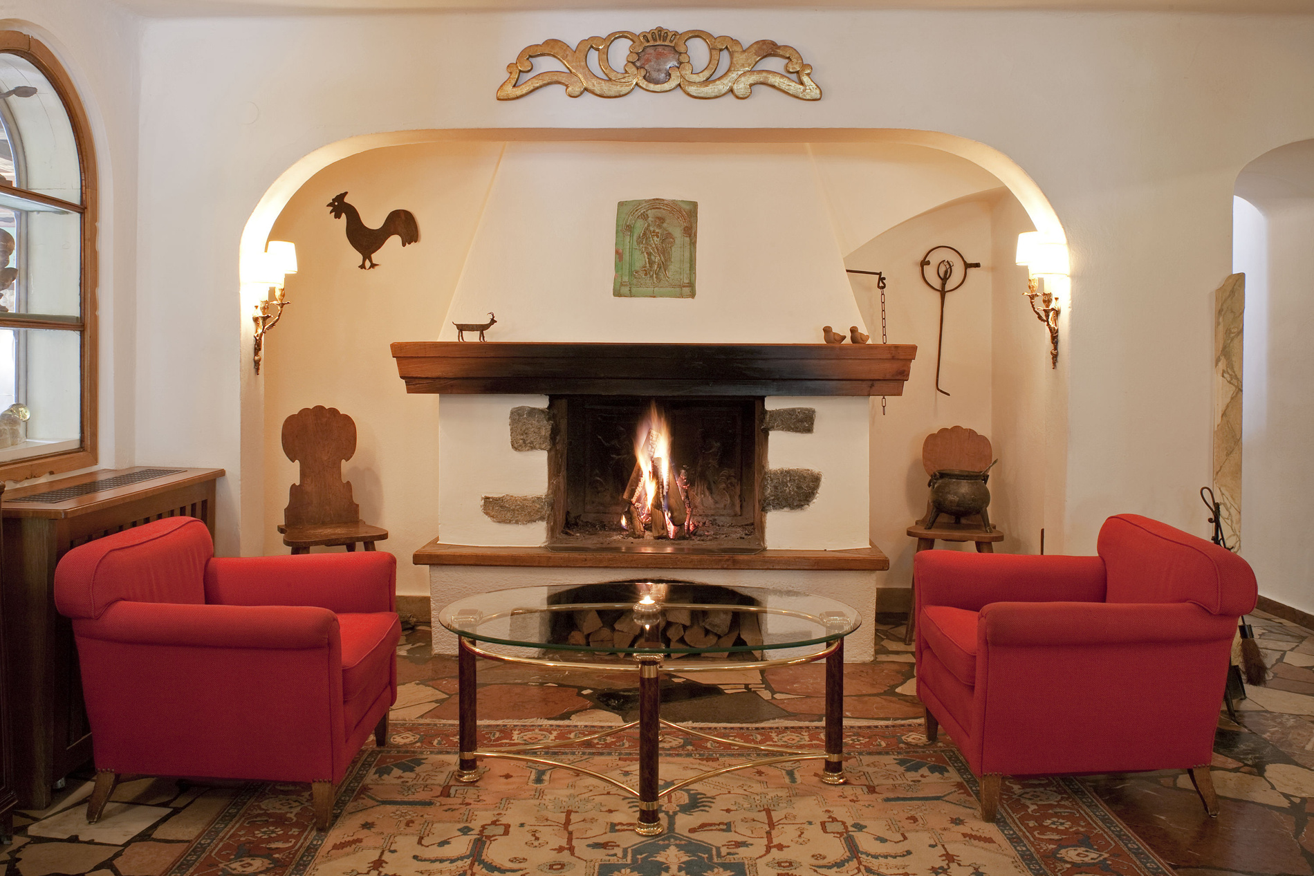 A cozy fireplace with a fire burning, flanked by red chairs and decorated with rustic art and ornaments.