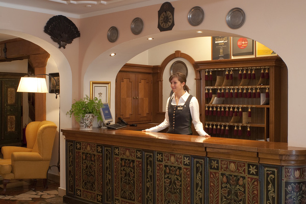 Eine Hotelrezeptionistin steht hinter einem verschnörkelten hölzernen Empfangstresen, hinter ihr hängen Zimmerschlüssel an der Wand.