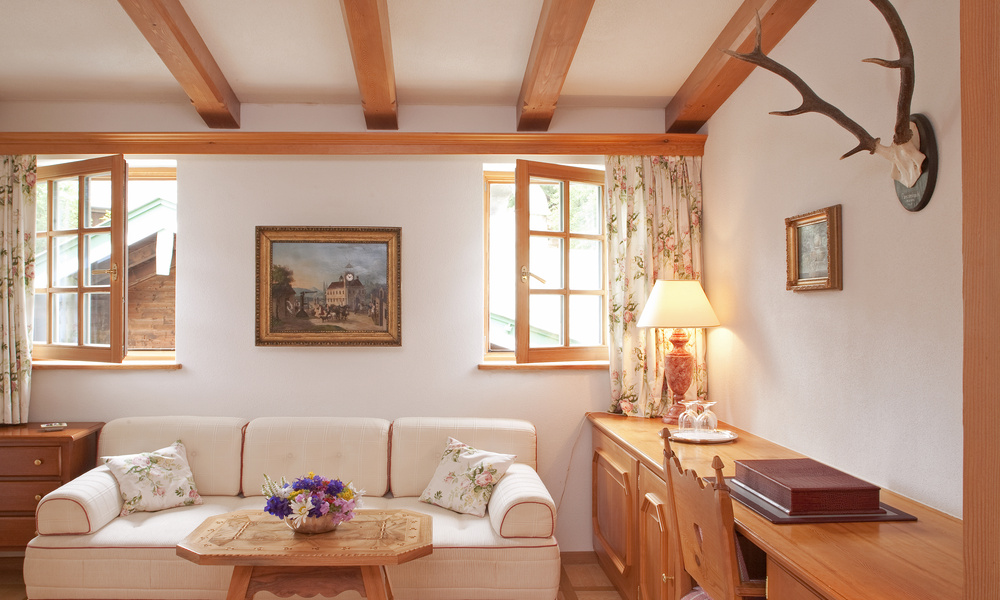 Gemütliches Wohnzimmer mit weißem Sofa, geblümten Vorhängen, Holzbalken, offenen Fenstern und einem Schreibtisch mit einer Lampe.