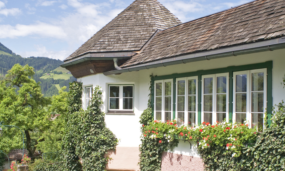 Ein Haus mit großen Fenstern, Efeu an den Wänden und roten Blumen, eingebettet in eine grüne, hügelige Landschaft.
