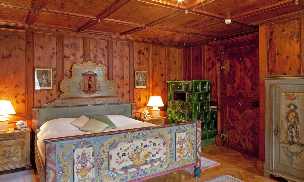 Gemütliches Holzzimmer mit bemalten Möbeln, grünem Kachelofen und zwei Lampen auf Nachttischen.